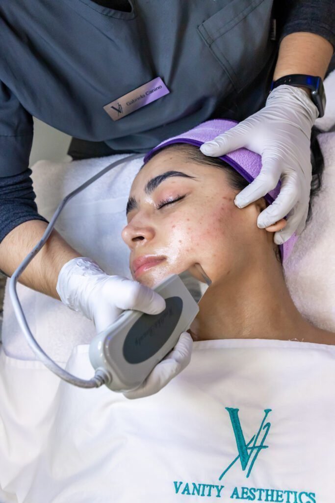 A woman relaxes with a red light therapy mask on her face, with a microdermabrasion tool and hydrating mask nearby, showcasing a combination approach to facial beauty treatment.