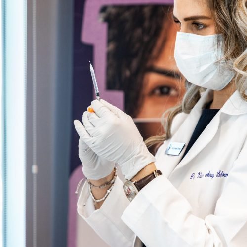 A woman receiving a laser treatment on her face at Vanitya Aesthetics. A technician in white gloves holds a device emitting a bright light against her cheek.